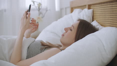 Woman-in-morning-sitting-in-bedroom-leaning-against-the-soft-leather-back-of-bed.-Write-messages-with-your-smartphone-use-apps-to-information